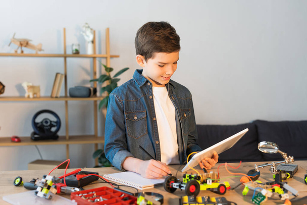αγόρι με ψηφιακή ταμπλέτα γράφοντας στο σημειωματάριο κοντά σε λεπτομέρειες του μοντέλου ρομποτικής στο τραπέζι - Φωτογραφία, εικόνα