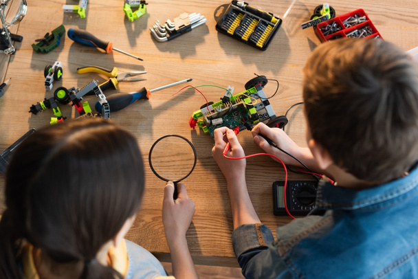 top view of boy with multimeter and girl with magnifying glass assembling robotics model at home - Foto, Imagem