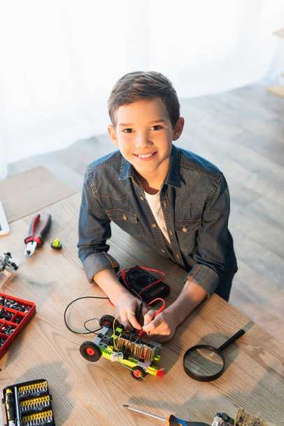 vysoký úhel pohledu usměvavého chlapce s multimetrem měřící napětí robotického modelu poblíž lupy  - Fotografie, Obrázek