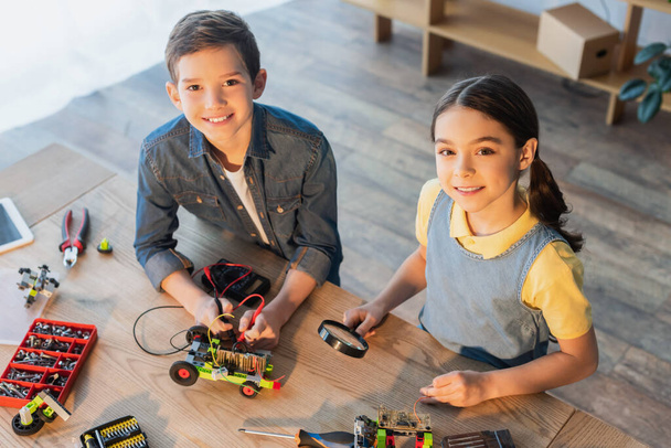 high angle view of kids with multimeter and magnifier assembling robotics model and smiling at camera - Photo, Image