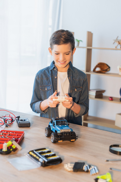 niño feliz sosteniendo el controlador remoto cerca del modelo de coche hecho a mano en la mesa en casa - Foto, imagen