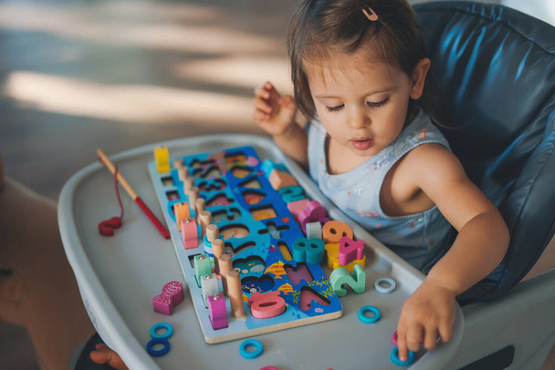 Shot of baby girl sitting on the high chair while learning and playing wooden toys at home. Infant in kindergarten. Colorful portrait. Baby development. - Φωτογραφία, εικόνα