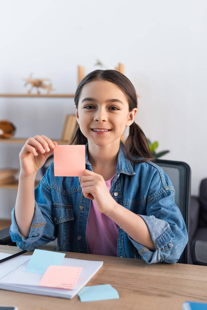 cheerful girl showing empty sticky note and looking at camera at home - Foto, Imagem