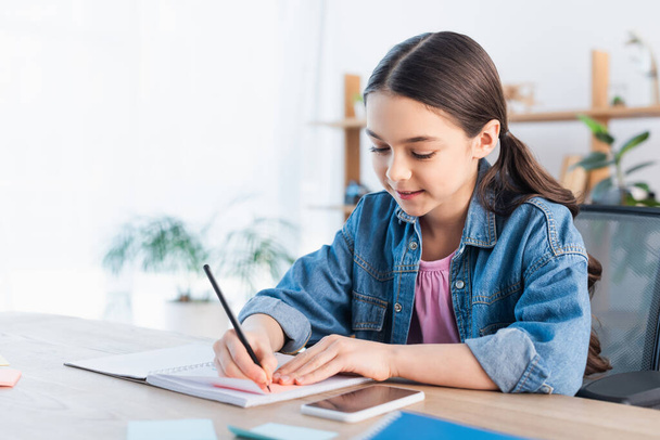 smiling brunette girl writing in notebook while doing homework near cellphone with blank screen - Photo, image