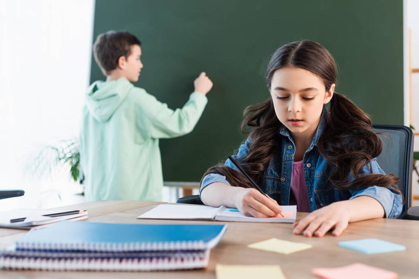 brunette schoolgirl writing in notebook near boy at chalkboard on background - Photo, image