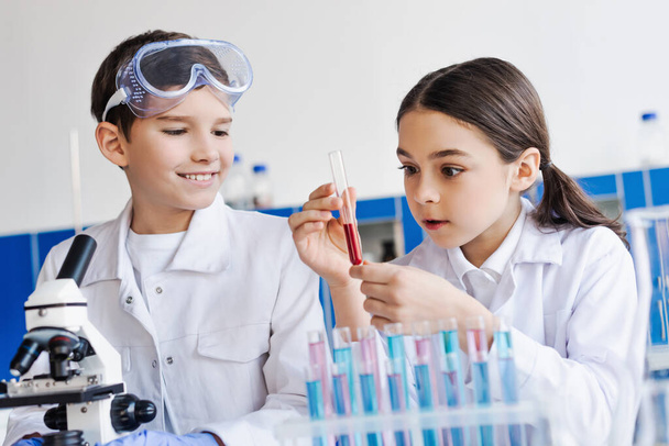 κατάπληκτος κορίτσι κρατώντας δοκιμαστικό σωλήνα με κόκκινο υγρό κοντά χαμογελαστό αγόρι και μικροσκόπιο στο εργαστήριο - Φωτογραφία, εικόνα