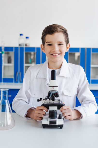 happy preteen kid in white coat looking at camera near microscope in laboratory - Foto, afbeelding