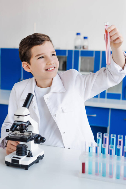 Glücklicher Junge blickt im Chemielabor auf Reagenzglas neben Mikroskop - Foto, Bild
