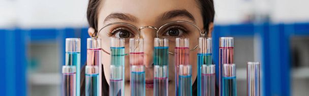 nastolatka dziewczyna w okularach patrząc na aparat w pobliżu probówek w laboratorium, baner - Zdjęcie, obraz