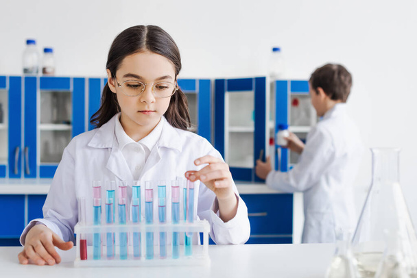 girl in white coat and eyeglasses near test tubes with liquid and boy on blurred background - Foto, imagen