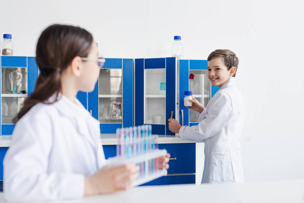 smiling boy in white coat holding jar with powder near girl with test tubes on blurred foreground - Photo, Image