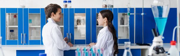 side view of kids in white coats looking at each other near locker in chemical lab, banner - 写真・画像