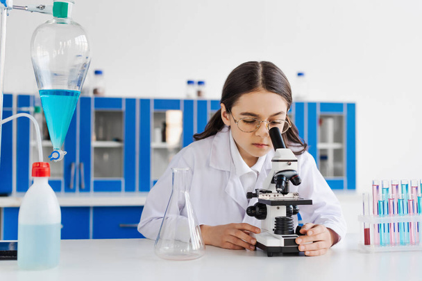 preteen girl in eyeglasses looking in microscope near test tubes and flasks - Fotoğraf, Görsel