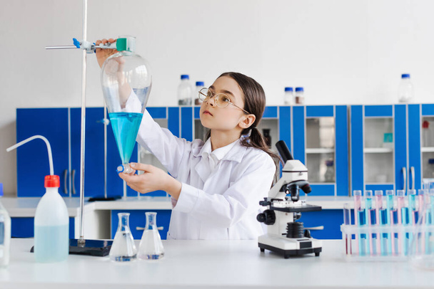 tiener meisje in bril en witte jas holding flacon in de buurt van microscoop en reageerbuizen - Foto, afbeelding