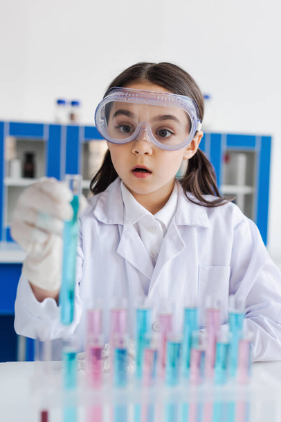 amazed girl in white coat and goggles looking at blurred test tube in lab - Foto, imagen