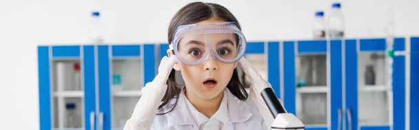 shocked girl in goggles touching head and looking at camera in chemical laboratory, banner - Photo, Image
