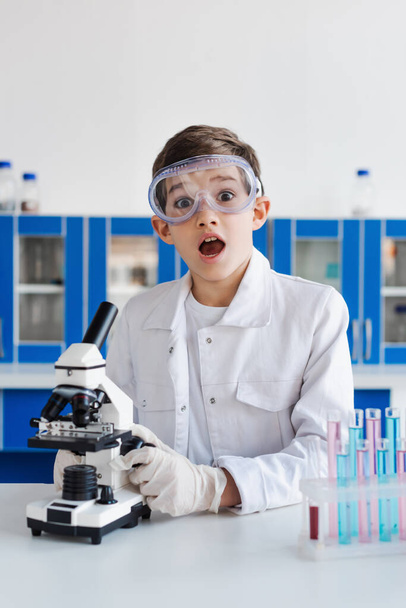 amazed boy in goggles looking at camera near microscope and test tubes in laboratory - Photo, Image