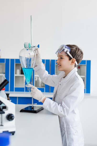 side view of smiling kid in latex gloves looking at flask with blue liquid near microscope - Foto, Imagem