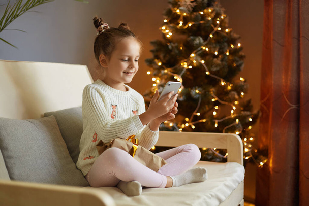 Image of joyful little girl with two hair buns wearing casual clothing sitting on sofa with mobile phone and taking selfie or having video call, congratulating friends with new year. - Foto, Imagen