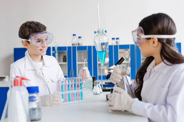 smiling boy in goggles looking at blurred girl near test tubes and flasks in lab - Photo, Image