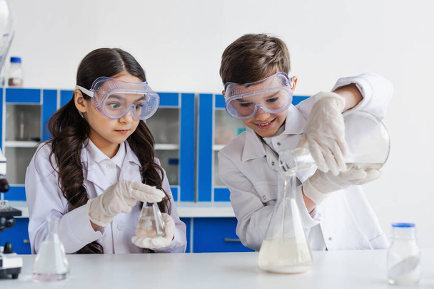 chica tensa en gafas mirando al niño sonriente vertiendo líquido en el frasco mientras hace experimento químico - Foto, Imagen