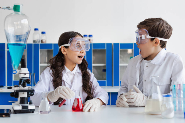 amazed girl in goggles looking at friend near flask with red liquid in chemical lab - Foto, afbeelding