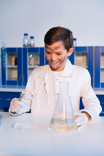 lachende jongen met vuil gezicht pincet in de buurt van petrischaal en kolf in lab - Foto, afbeelding