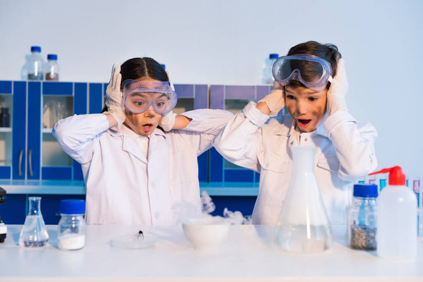 verblüffte Kinder mit dreckigen Gesichtern, die Köpfe berühren, während sie dampfende Schüssel im Chemielabor betrachten - Foto, Bild