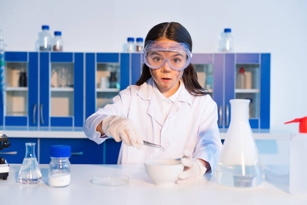 surprised girl with dirty face holding tweezers near bowl and flask with steam in lab - Photo, Image