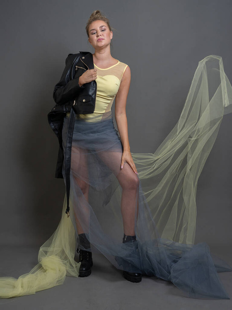 Fashion blonde woman in black eco leather boots, jacket, transparent tulle skirt, studio shot on gray background, vertical photo, full length portrait - 写真・画像