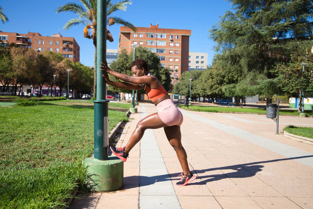 トレーニング前に公園でストレッチ彫刻体を持つ若くて美しいアフリカ系アメリカ人女性。彼女はオレンジの上とオレンジのレギンスを着ています。スポーツと健康の概念. - 写真・画像