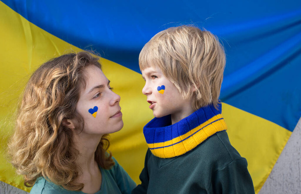 мальчик и молодая женщина с раскрашенным желто-синим сердцем на щеках на фоне украинского флага. Семья, единство, поддержка. Дети против войны. призыв к мировому сообществу о помощи украинцам - Фото, изображение