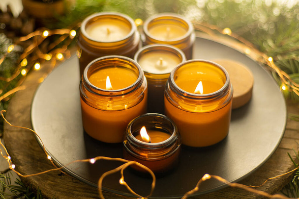 Soy candles burn in glass jars. Comfort at home. Candle in a brown jar. Scent and light. Scented handmade candle. Aroma therapy. Christmas tree and winter mood. Cozy decor. Festive garland decoration - Φωτογραφία, εικόνα