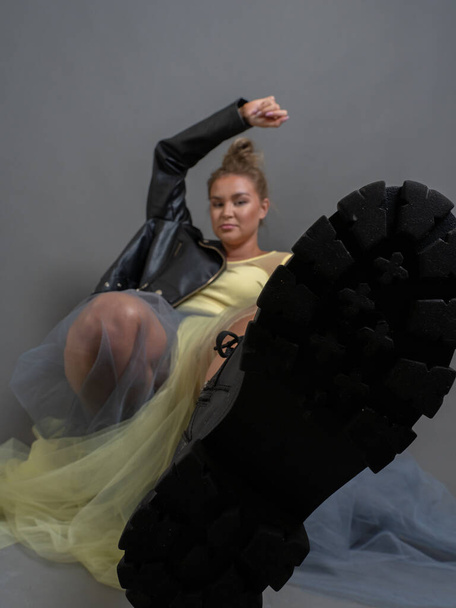 Fashionable blonde woman in black eco-leather boots, jacket, transparent tulle skirt, studio shot on a gray background, vertical photo, shoes in focus in the foreground - Foto, Bild