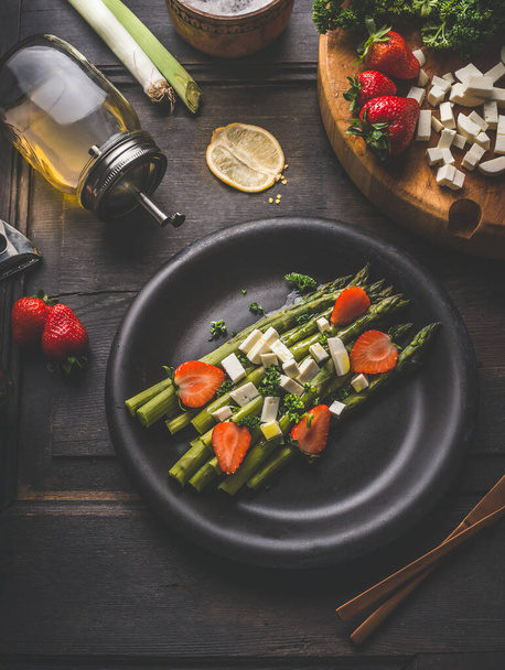 Tasty asparagus salad topped with strawberries and fresh cheese cubes served in black plate on dark rustic table background with ingredients. Top view. Healthy food. Home cooking and eating - Foto, Imagen