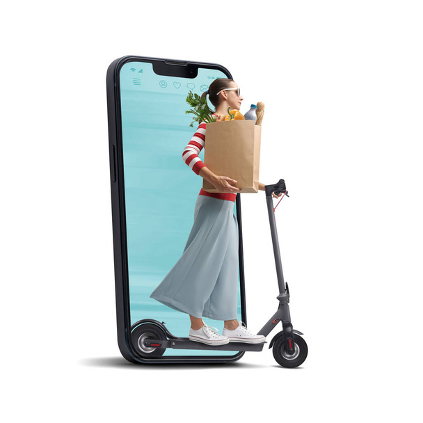 Woman holding a paper bag with groceries and riding an electric scooter, she is coming out from a smartphone screen, online grocery shopping concept - Photo, Image