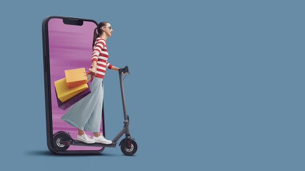 Woman holding many shopping bags and riding an electric scooter, she is coming out from a smartphone screen, online shopping and sales concept - 写真・画像