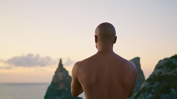 Sterke man die yoga beoefent mediterend bij zonsondergang oceaanklif zicht. Kalm als een man die zich concentreert op het maken van namaste gebaar. Onbekende yogi die spirituele oefening uitvoert op zoek naar innerlijke harmonie achteraanzicht - Foto, afbeelding