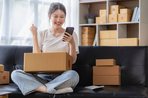Asian woman sitting on sofa at home working freelance using laptop with box, successful Asian woman with raised hand, online marketing packaging and delivery box, SME concept - Photo, Image
