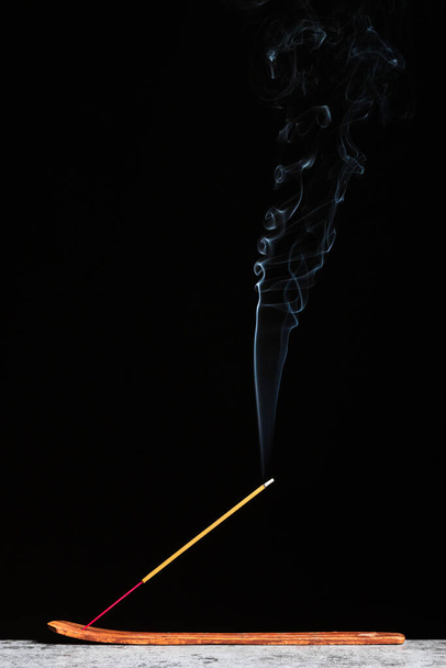 Burning smoking incense stick stands on a wooden stand isolated on a black background. - Valokuva, kuva