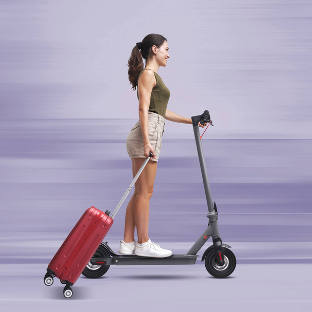 Tourist woman riding a fast electric scooter and carrying a trolley bag, sustainable mobility concept - Fotó, kép