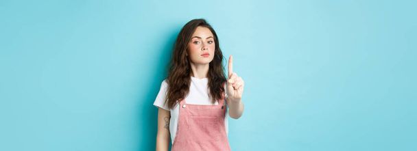 Rule number one. Serious young beautiful girl shaking finger in disapproval, scolding bad decision, make block or taboo gesture, standing against blue background. - Photo, Image