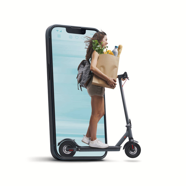Woman holding a paper bag with groceries and riding an electric scooter, she is coming out from a smartphone screen, online grocery shopping concept - Foto, Bild