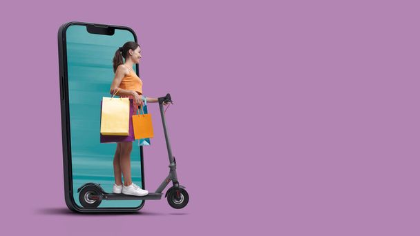 Happy woman holding shopping bags and riding a scooter, she is coming out from a smartphone screen, online shopping and sales concept - Photo, Image