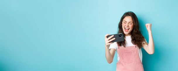 Cheerful cute girl winning in online video game on smartphone, making fist pump and shouting yes with joy, standing over blue background and triumphing. - Photo, Image