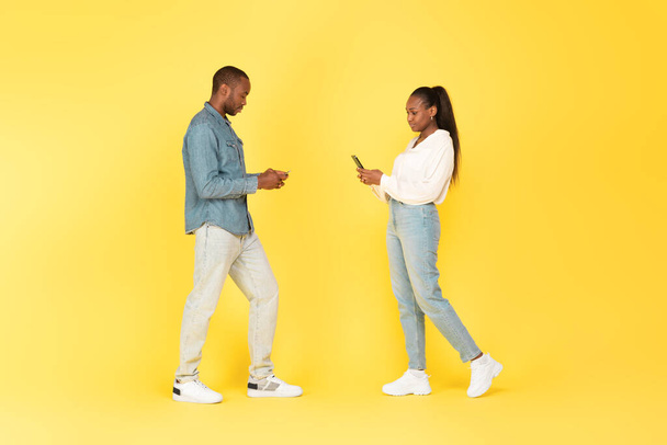 Schwarzer Mann und Frau mit Smartphones, die im Internet surfen und SMS schreiben, stehend über gelbem Studiohintergrund, Aufnahme in voller Länge. Neue Werbung für mobile Anwendungen - Foto, Bild