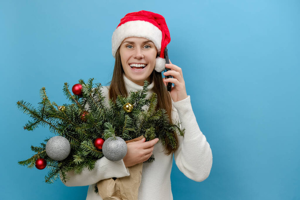 Happy beautiful young female in Christmas hat holding bouquet of spruce branches talk speak on mobile cell phone conducting pleasant conversation, posing over blue color background wall in studio - Foto, Imagen