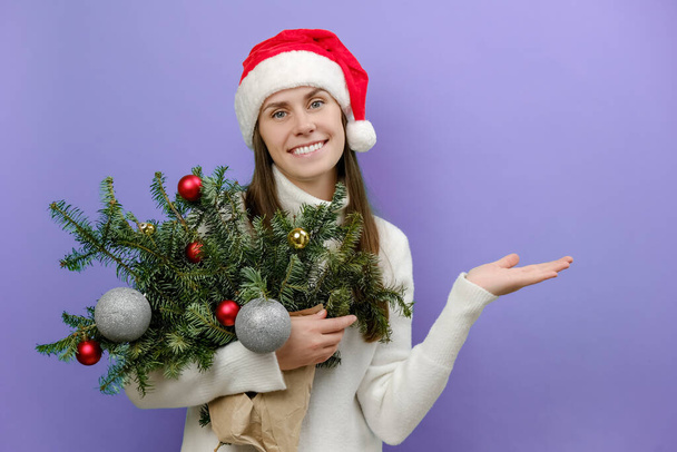 Pozitív fiatal nő a karácsonyi piros kalap kezében csokor lucfenyő ágak bemutató tenyérrel, pózol lila színű háttér stúdió fénymásolás. Boldog új évet ünneplés koncepció - Fotó, kép