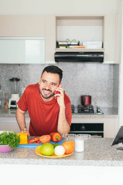 Мужчина разговаривает по мобильному телефону, когда готовит завтрак дома - Фото, изображение