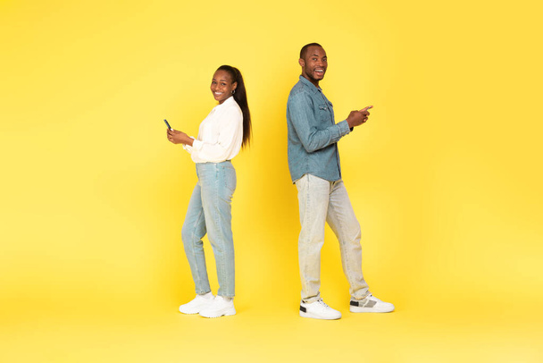 Allegro nero coppia utilizzando i telefoni cellulari sms in piedi back to back su sfondo giallo Studio. Comunicazione mobile, pubblicità delle applicazioni. Colpo a figura intera - Foto, immagini